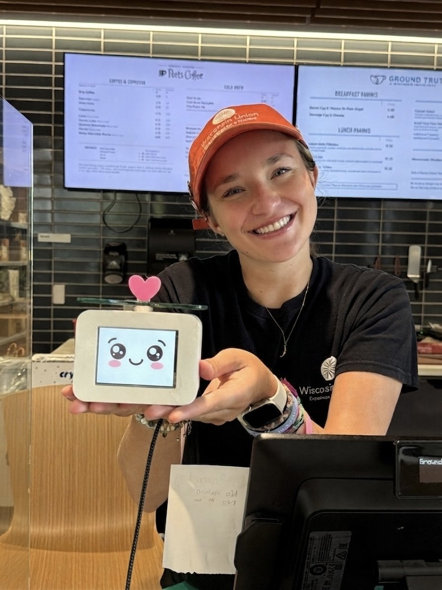 A barista at a campus coffee shop smiling and holding the Cubot prototype, which displays a kawaii-style blushing face