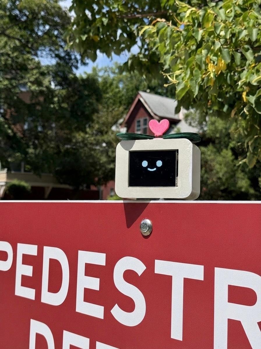 Cubot prototype mounted on top of a pedestrian sign outdoors, displaying a smiling face, with a house and trees in the background