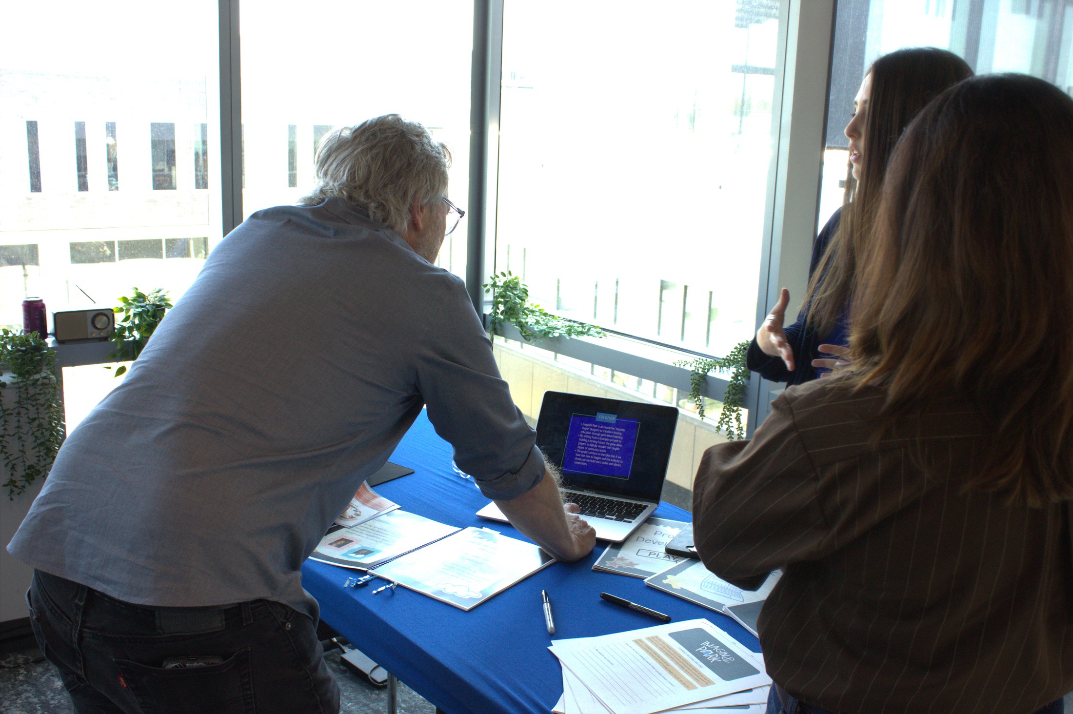 A visitor leaning over the exhibit table to look at the laptop screen while two team members explain the project
