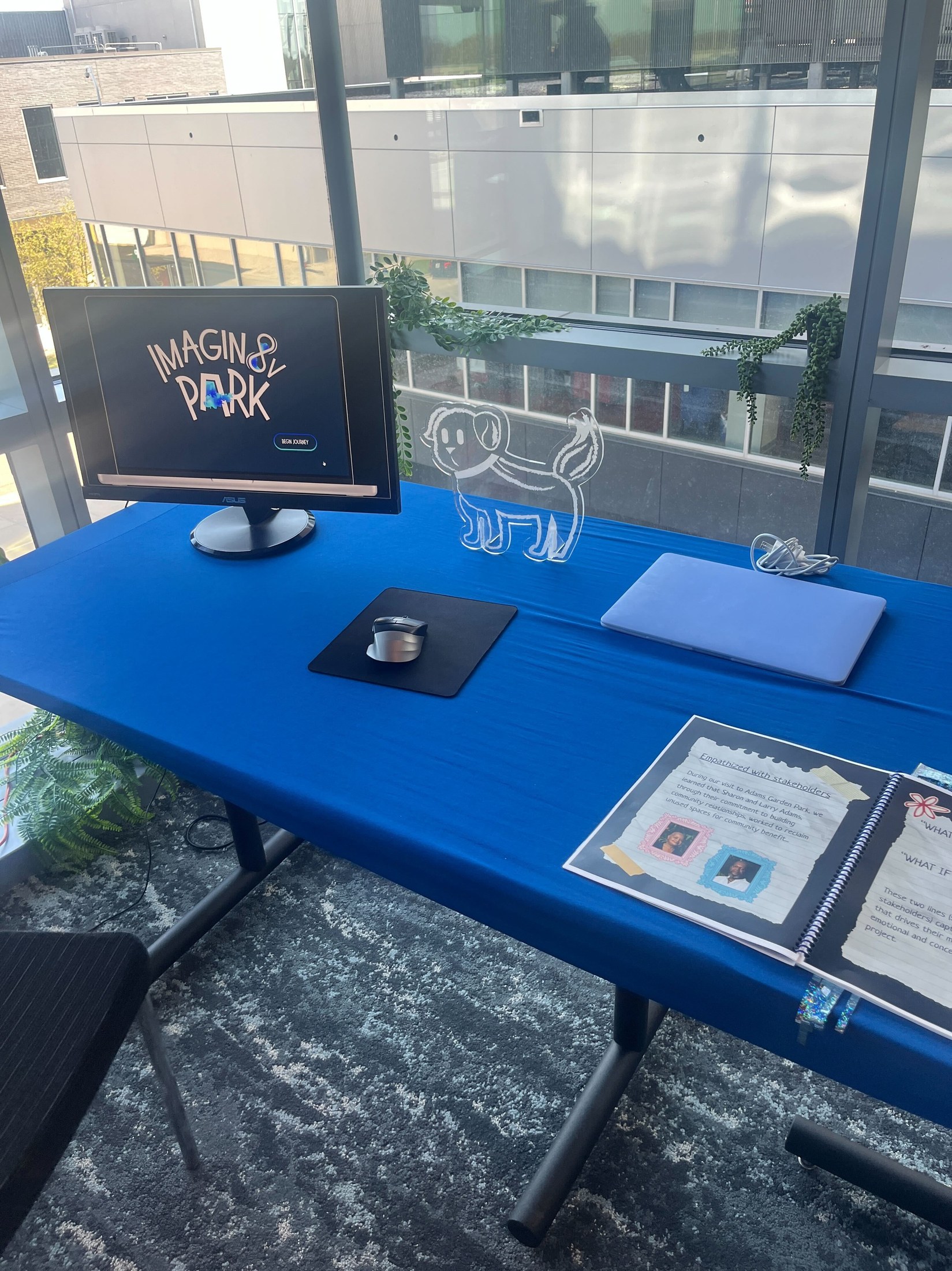 The exhibit kiosk: a monitor showing the Imagin8V Park Begin Journey screen, a laser-cut acrylic silhouette of Terry the ghost dog, our printed process book, a laptop, and a worksheet introducing stakeholders Sharon and Larry Adams of Adams Garden Park