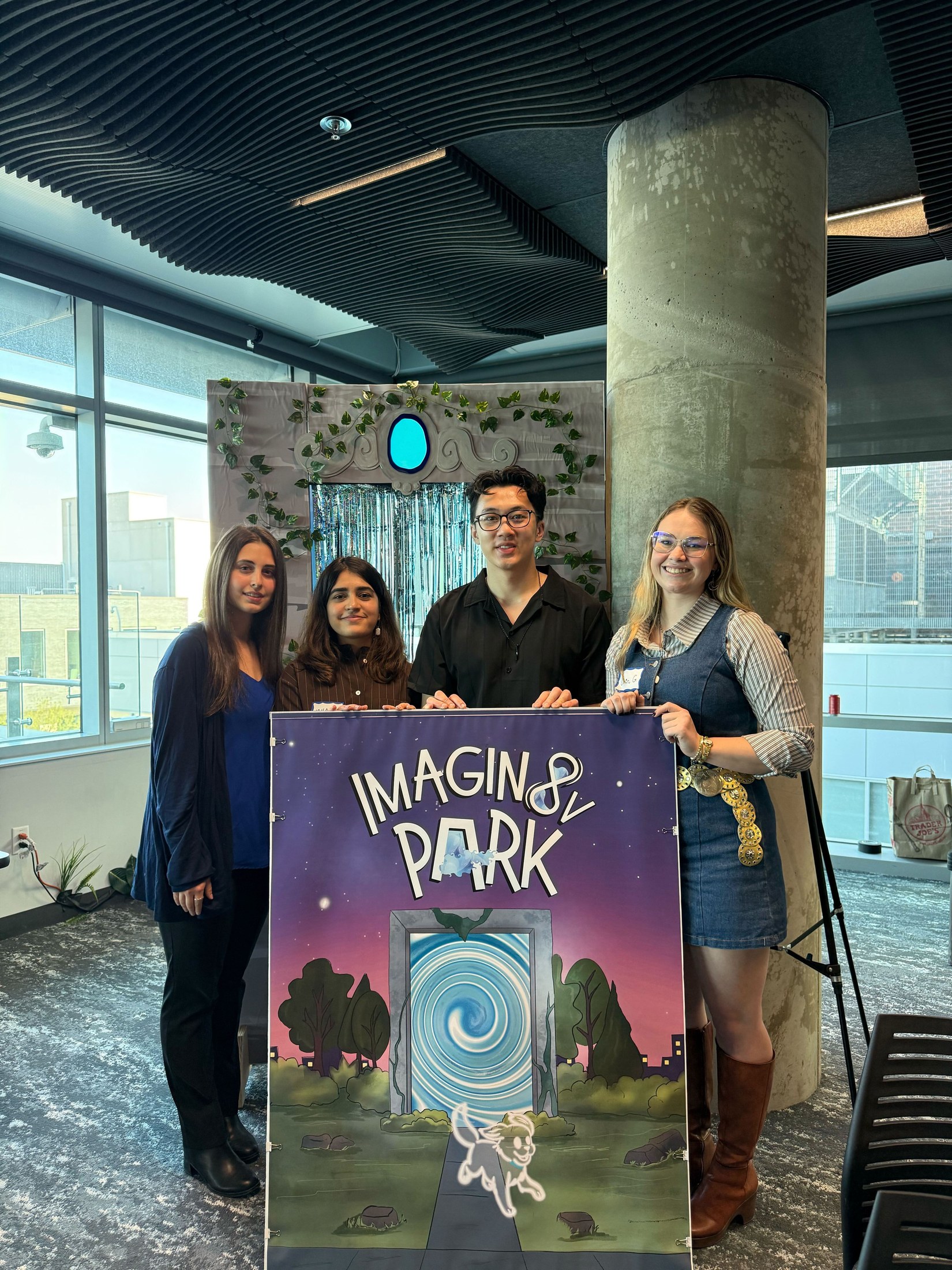 Four team members standing behind a physical cardboard portal cutout of the Imagin8V Park doorway, holding a sign with the project logo